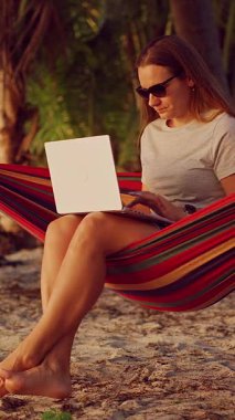 Dikey video. Sıcak günbatımı kumlu sahili aydınlatırken bir kadın, tropik palmiyelerin altında rahat bir şekilde otururken dizüstü bilgisayarda çalışır.