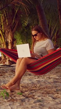 Dikey video. Bir kadın, sıcak gün batımı ışığı olan tropik palmiyelerin altında rahat bir şekilde oturup rahat bir şekilde dizüstü bilgisayarda çalışarak rahat bir çalışma ortamı yaratıyor.