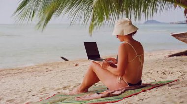 Kadın, sakin okyanusa bakan dizüstü bilgisayarla uzaktan çalışmaya devam ederken Palm Leaves 'in altında rahat bir şekilde oturuyor.