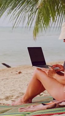 Dikey video. Kadın, sakin okyanusa bakan dizüstü bilgisayarla uzaktan çalışmaya devam ederken Palm Leaves 'in altında rahat bir şekilde oturuyor.