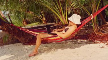Hamak altında yumuşak tropikal ışık ve kumlu zemin ile uzanan bir kadın dizüstü bilgisayarında yazı yazıyor.