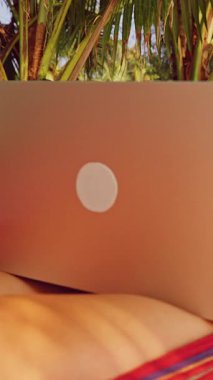 Dikey video. Sunny Beach 'te, Palm Trees' in altında dizüstü bilgisayar kullanan odaklanmış bir kadın modern uzaktan çalışma ve seyahat özgürlüğü yaşam tarzını temsil ediyor.