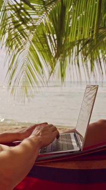 Dikey video. Sıcak Tropikal Gün 'de Hamakta Yüzü Parlayan Deniz Altında Çalışan Kadın Dizüstü Bilgisayarda Çalışıyor