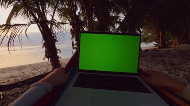 Yeşil ekranlı dizüstü bilgisayarın ilk perspektifi tropik plajın yakınındaki hamakta dinleniyor, Palm Trees ve Sessiz Okyanus Altın Saati Uzaktan Çalışma ve Seyahat Yaşam Tarzı