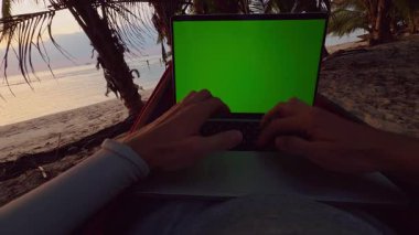 Güneş batarken hamakta uzanan tropikal plajda dizüstü bilgisayarla yazı yazan ilk insan görüntüsü Palm Trees ve sakin deniz uzaktan çalışma ve dijital göçebe yaşam tarzı yaratıyor.