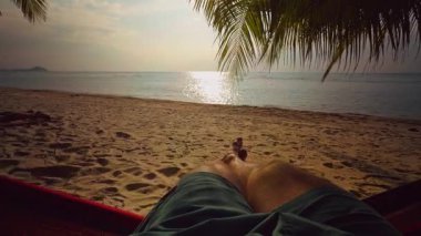 Kum tropikal plajında palmiye yaprakları ve gün batımında ışıldayan denizle dinlenen çıplak bacakların ilk görüntüsü sakin bir tatil ve yavaş bir yaşam havası yaratır.