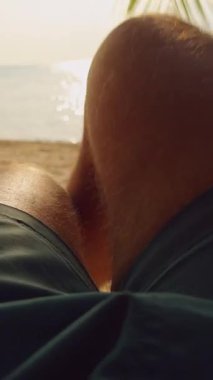 Dikey video. Günbatımında sakin bir deniz ve tropikal plaja bakan hamağın gevşek bacaklarının ilk insan görüntüsü dinlenme ve tatil yaşam tarzını vurguluyor.
