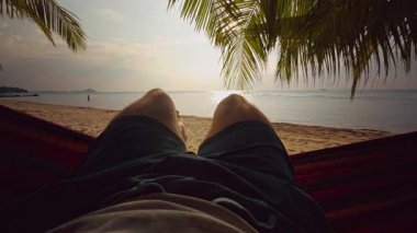 Hamakta kesişen ilk kişi Palm Trees 'in altında Tropikal plaja ve sessiz okyanusa bakan altın gün batımında kaygısız yolculuk havasına neden olan bacaklar.