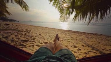 Hamaktaki ilk insan görüntüsü Tropikal plaja doğru uzanan palmiye yaprakları ve gün batımında ışıldayan denizle özgürlük ve rahat tatil yaşam tarzını gösteren