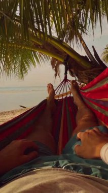 Dikey video. Hamakta sallanan insanın ilk perspektifi, Ilık günbatımında Parlayan Denize ve Palmiye dallarına doğru uzanan Bacakları ile Sakin Yolculuk Ruh halini gösteren