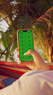 Dikey video. Tropikal Palm Garden 'da Yeşil Ekran ile Hamak Smartphone Besleyen Kadın, Sosyal Medya Taraması ve Mobil Teknoloji Eğlencesi İlk Kişi Görünümü