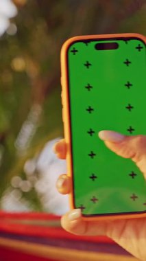 Dikey video. Hamakta Sallanan Kadın Akıllı Telefon Tropik Palm Garden 'da Yeşil Ekran ile Kaydırırken, Sosyal Medya Taraması ve Rahat Mobil Yaşam Taraması Gösteren İlk Kişi Görünümü