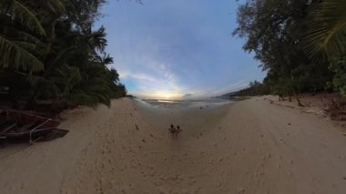 Geniş Tropikal Kıyı Boyunca Yürüyen Bir Çift Küresel Bir Panorama Efektiyle Canlı Bir Günbatımına Doğru Uzun Palmiyeler İki Tarafta da Yükseliyor ve Yumuşak Akşam Işığı Huzurlu Bir Romantik Atmosfer Oluşturuyor
