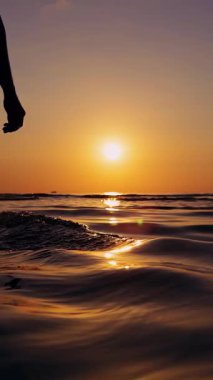 Dikey video. Gün batımında sığ denizde ilerleyen kişinin düşük açılı görüntüsü Altın gökyüzüne karşı karanlık siluet ve yavaş hareketler sakin bir yolculuk ve iç hareket yaratıyor.