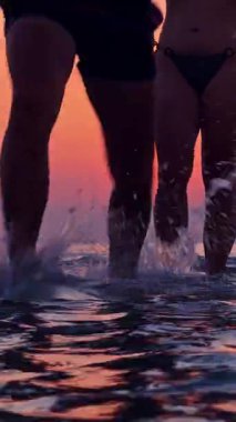 Dikey video. Gün batımında kadın onun yanında suyla hareket ederken, erkeğin sığ denizde ilerleyişinin düşük açılı görüntüsü sıcak gökyüzüne sıçrıyor ve Dinamik Yaz Hareketi paylaşılıyor.