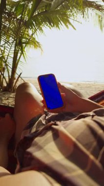Dikey video. Hamakta Dinlenen Kadın Tropikal Plaj Tutucu Akıllı Telefon Parlak Mavi Kroma Anahtar Ekranlı Palmiye Ağaçları ve Sıcak Güneş Işığıyla çevrili Rahat Seyahat Yaşam Tarzı