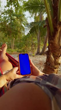 Dikey video. Palm Trees 'in Altında Dinlenen Kadın Tropikal Plajda Mavi Krom Anahtar Ekranlı Akıllı Telefon Kullanıyor