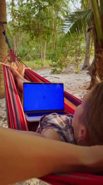 Dikey video. Hamakta Yatan Kadın Parlak Mavi Krom Anahtarlı Laptop kullanarak Tropikal Plaj 'da Palmiye Ağaçlarının Altında Uzaktan Çalışma Ortamı Oluşturuyor