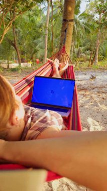 Dikey video. Hamakta Rahat Kadın Dizüstü bilgisayarlı Mavi Krom Anahtar Ekranlı Mockup Tropikal Palmiye Ormanı ve Kumsal Sahili ile çevrili