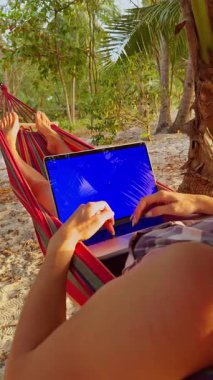 Dikey video. Kadın Eller Mavi Krom Anahtarlı Dizüstü Bilgisayarı Kullanırken Hamak Altında Tropik Plaj Gösterimi Dengesi 'nde Palmiye Ağaçları Altında Dinleniyor