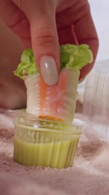 Dikey video. Taze bahar rulosu sosa batırıldı. Plaj havlusunda kadın eli tarafından tutuldu. Arka planda denizle birlikte, iştah açıcı sağlıklı bir aperatif anı yarattı.