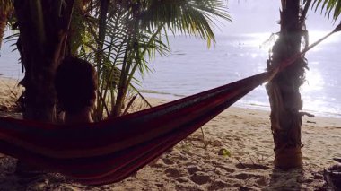 Kumlu tropik bir plajda palmiye ağaçları arasında dinlenen adam güneş ışığının yansımasıyla sakin bir denize bakıyor ve yavaş ada rahatlama havası yaratıyor.