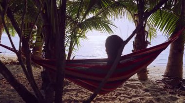 Palmiye yapraklarının altında bir hamakta uzanan adam yumuşak gölgeler ve ılık ışıkla parlak okyanusa bakıyor ve huzurlu tropikal plaj atmosferi yaratıyor.