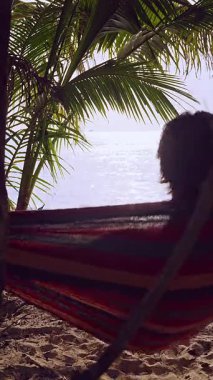 Dikey video. Palmiye yapraklarının altında bir hamakta uzanan adam yumuşak gölgeler ve ılık ışıkla parlak okyanusa bakıyor ve huzurlu tropikal plaj atmosferi yaratıyor.