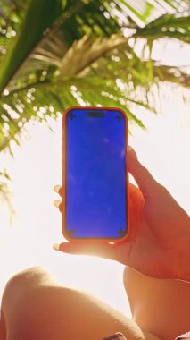 Dikey video. Hamakta Bir Kadın Tarafından Mavi Krom Ekranlı Akıllı Telefon Tropikal Doğa Resmetme Mobil Uygulama ve Boş Zaman Sunumu
