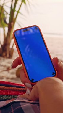 Dikey video. Palmiye Ağaçları Arasındaki Hamakta Dinlenen Kadın tarafından kullanılan Mavi Model Ekranlı Akıllı Telefon Dijital Özgürlük ve Mobil Yaşam Tarzını Temsil Ediyor