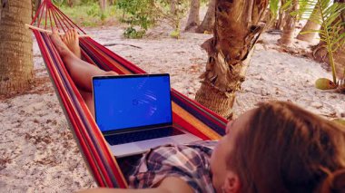 Hamakta yatan genç kadın, kumlu sahildeki palmiye ağacının altında mavi renk ekranlı dizüstü bilgisayarı, tropikal uzaktan kumandalı çalışma sahnesi, dijital içerik ve seyahat yaşam tarzı için cihaz modeli