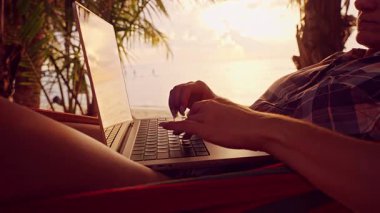Gündoğumunda hamakta yatarken dizüstü bilgisayarda yazan bir kadın, tropik plaj uzaktan çalışma sahnesi, okyanus manzaralı rahat dijital göçebe yaşam tarzı.