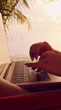 Dikey video. Gündoğumunda hamakta yatarken dizüstü bilgisayarda yazan bir kadın, tropik plaj uzaktan çalışma sahnesi, okyanus manzaralı rahat dijital göçebe yaşam tarzı.