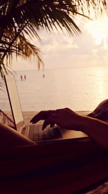 Dikey video. Hamakta dinlenen genç kadın tropikal plaj gündoğumu, dingin okyanus arka planı, dijital göçebe iş hayatı dengesi üzerinde çalışırken