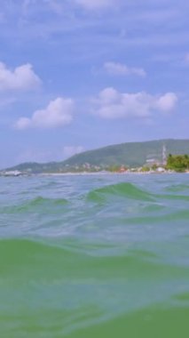 Dikey video. Tropikal deniz yüzeyi manzarası, yumuşak dalgalar palmiye plajına doğru yuvarlanıyor, ufukta yavaş hareket eden su dokusu, mavi gökyüzü ve uzak tepe, huzurlu yaz yolculuğu havası yaratıyor.