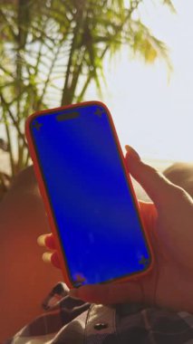 Dikey video. Mavi ekran ile el ele tutuşan akıllı telefonun yakın görüntüsü tropik plajın yanındaki hamakta, sıcak güneş ışığı ve sakin sahil atmosferinde yatarken