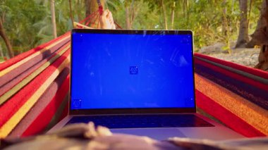 Tropikal ormanlardaki hamakta dinlenen boş mavi ekran dizüstü bilgisayarlı, ayakları görünen, uzaktan kumandalı ve rahat dijital yaşam tarzı olan ilk kişi.