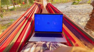 Hamakta dizüstü bilgisayarı bacaklarının üzerinde yatan biri, mavi ekran görünür, tropikal zemin ve etraftaki ağaçlar, rahat uzaktan çalışma ve yavaş yaşam ortamı