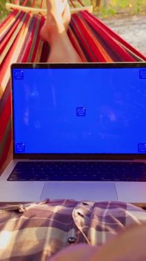 Dikey video. Hamakta dizüstü bilgisayarı bacaklarının üzerinde yatan biri, mavi ekran görünür, tropikal zemin ve etraftaki ağaçlar, rahat uzaktan çalışma ve yavaş yaşam ortamı