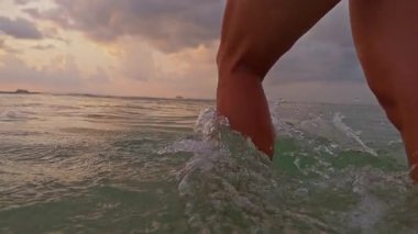 Gün batımında sığ denizde yürüyen kadın, bacağının etrafında hafif dalgalar, dramatik bulutlu gökyüzü ve sıcak ufuk ışığı, yavaş çekim deniz kenarı özgürlüğü