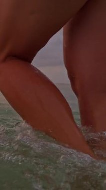 Dikey video. Gün batımında sığ denizde yürüyen kadın, bacağının etrafında hafif dalgalar, dramatik bulutlu gökyüzü ve sıcak ufuk ışığı, yavaş çekim deniz kenarı özgürlüğü