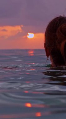 Vertical video. Woman resting in open sea while watching sunset, low waterline view with soft wave and glowing sky, slow motion tranquil evening atmosphere