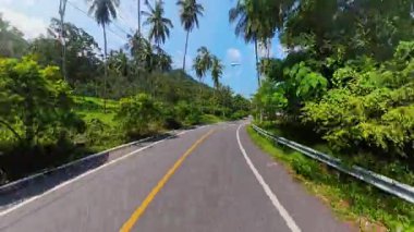 Boş Tropikal asfalt yolu Palmiye ağaçları ve açık mavi gökyüzünün altındaki yeşil tepeler arasında uzanır Huzurlu bir yolculuk ve ada manzarasını gösterir