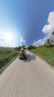 Dikey video. Tozlu kırsal yolda, palmiye ağacı ve parlak gökyüzü ile motosiklet süren çift, geniş açı sahnesinde Tropikal Seyahat