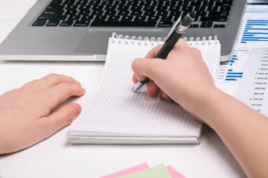 Masadaki kadın bir deftere yazıyor. Boş bir defterin üzerinde elinde kalem tutan bir adam. Bilgisayarlı ve mali tablolu ofis masası.