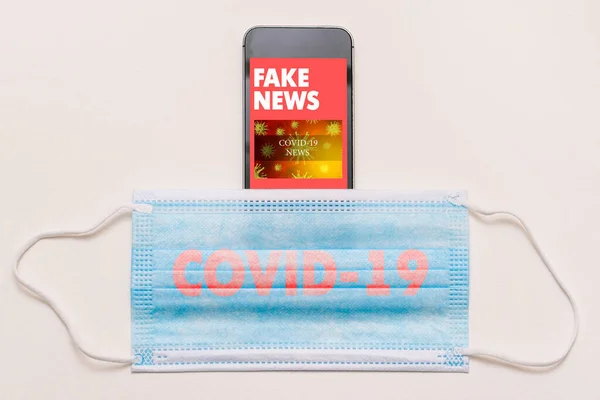 Fake news concept on coronavirus theme. Mobile phone and medical mask with COVID-19 text. News feed on smartphone screen with fake articles.