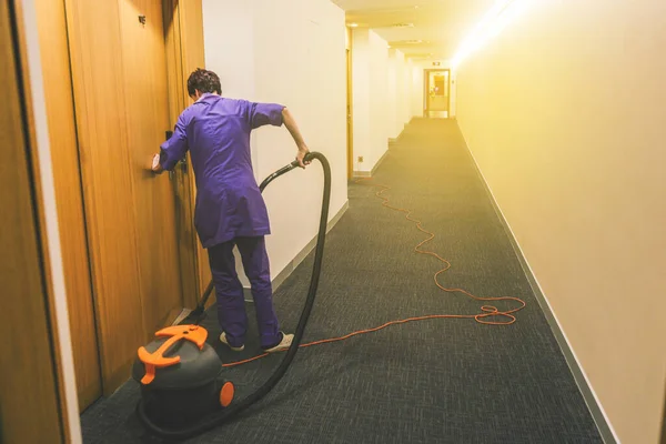 Η γυναίκα καθαρίζει το δωμάτιο του ξενοδοχείου. Υπηρεσίες καθαρισμού ξενοδοχείων. Δουλεύοντας ως υπηρέτρια στο ξενοδοχείο. Μακρύ διάδρομο ξενοδοχείου με ξύλινες πόρτες — Φωτογραφία Αρχείου