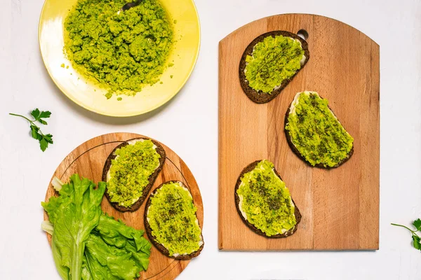 Sandwiches with avocado on a wooden board. Ingredients for making toast for breakfast or dinner. Yellow plate with mashed avocado, spices, salad on a white plate. Top view, flat lay.