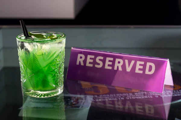 A glass with a green alcoholic drink stands on a glass table, next to the plate reserved, with a cocktail tube