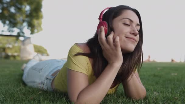 Молода іспаномовна жінка слухає музику з навушниками, відпочиваючи на зеленій траві в парку — стокове відео
